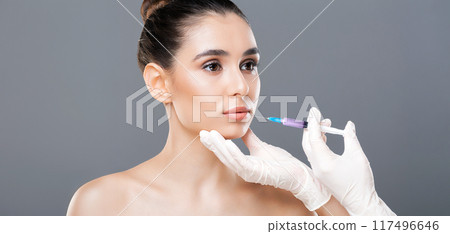 A woman is receiving a dermal filler injection administered by a professional. She sits calmly as a gloved hand holds the syringe near her face, emphasizing a meticulous and sanitary procedure. A woman is receiving a dermal filler injection administered by a professional. She sits calmly as a gloved hand holds the syringe near her face, emphasizing a meticulous and sanitary procedure. 117496646
