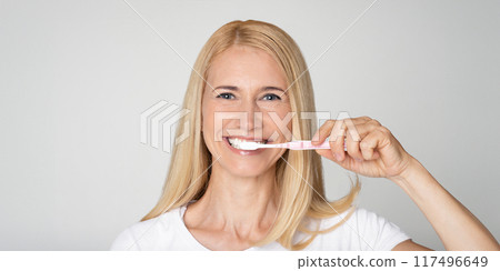 A blonde woman with a white shirt brushes her teeth with a pink toothbrush and white toothpaste in front of a plain white background. A blonde woman with a white shirt brushes her teeth with a pink toothbrush and white toothpaste in front of a plain white background. 117496649