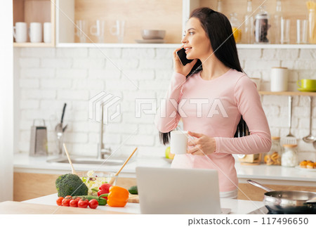 A young woman dressed in a pink long-sleeve shirt is standing in a bright, modern kitchen in the morning. She holds a white mug and is actively engaged in a phone conversation 117496650