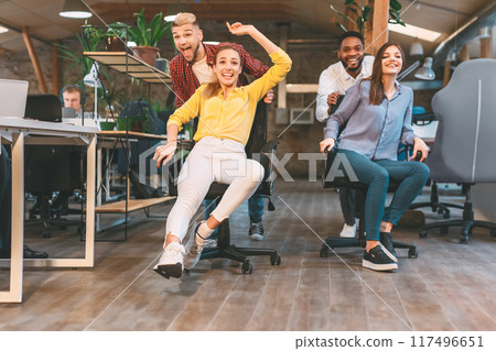 Time for break. Joyful colleagues having fun at workplace, making office chair race in coworking space Time for break. Joyful colleagues having fun at workplace, making office chair race in coworking space 117496651