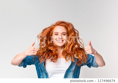 A Caucasian woman with vibrant red hair, wearing a casual outfit, is smiling and making a thumbs up gesture with her right hand towards the camera. A Caucasian woman with vibrant red hair, wearing a casual outfit, is smiling and making a thumbs up gesture with her right hand towards the camera. 117496674
