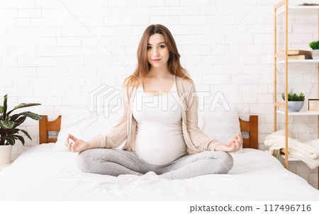 A pregnant woman is sitting cross-legged on a bed, practicing meditation. She has her hands resting on her belly, eyes closed in concentration. 117496716