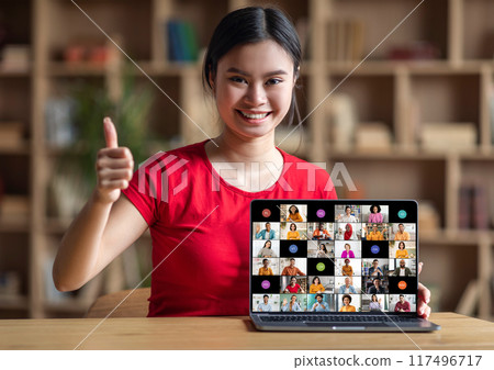 A young woman smiles and gives a thumbs up while sitting at her desk and participating in an online meeting on her laptop computer. The meeting is displayed on the screen 117496717