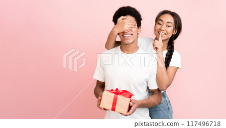 A cheerful African American couple engaged in a playful surprise gift exchange. The girl covers the guy eyes while holding a wrapped present that adds to the excitement of the moment. 117496718