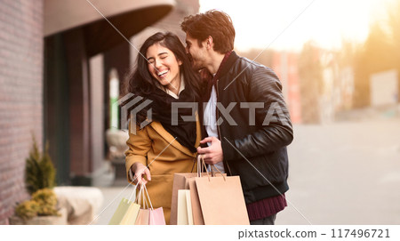 A man and woman are standing in front of a building, holding shopping bags. 117496721