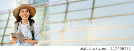 Glad young european woman student in hat with backpack, use tablet, look at empty space enjoy, trip on summer on station, work, sun flare. App for tourist travel, blog, ad and offer, copy space 117496731