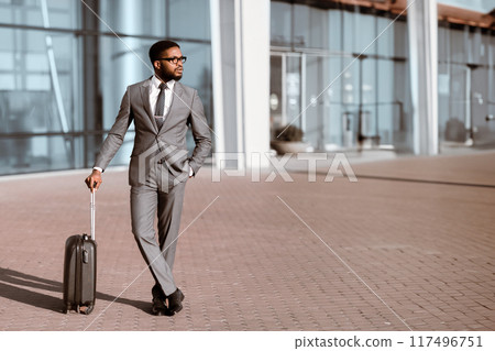 Black man executive is walking with luggage outside an airport terminal. The atmosphere is busy and professional, capturing the essence of corporate travel in a modern environment. Black man executive is walking with luggage outside an airport terminal. The atmosphere is busy and professional, capturing the essence of corporate travel in a modern environment. 117496751