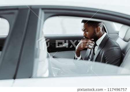 African American thoughtful businessman with a beard, wearing a suit and tie, sits in the back seat of a modern sedan. His hand is resting on his chin, and his gaze is directed down 117496757