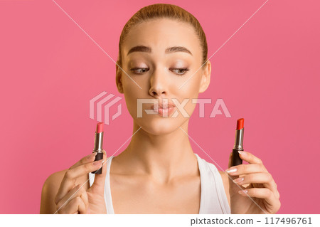 A young woman with blonde hair and a white shirt stands in front of a pink background. She is looking at the camera with a thoughtful expression and holding two lipsticks 117496761