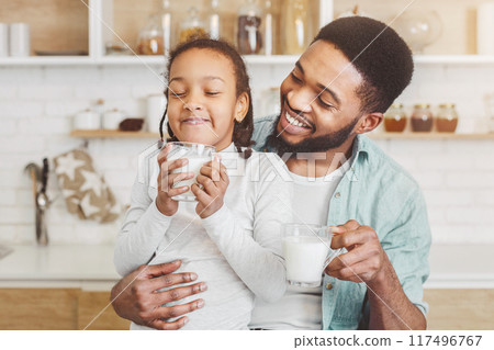 African father encouraging his sweet daughter to drink milk, kitchen background, free space African father encouraging his sweet daughter to drink milk, kitchen background, free space 117496767