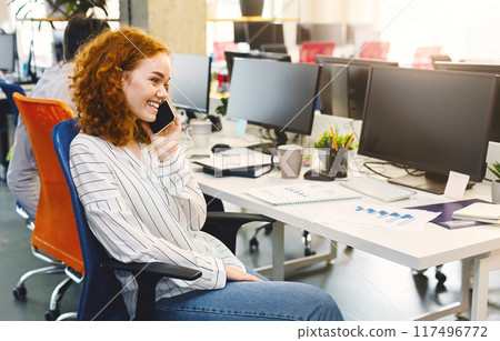 A woman sitting at a desk, engaged in conversation on her cell phone. She appears focused and attentive while speaking. The desk is neat and organized, with a computer and stationery on it. A woman sitting at a desk, engaged in conversation on her cell phone. She appears focused and attentive while speaking. The desk is neat and organized, with a computer and stationery on it. 117496772