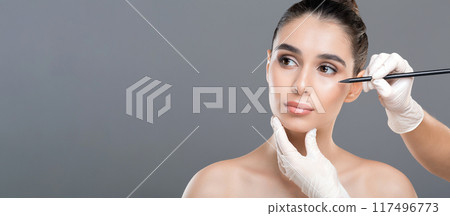A young woman is receiving a cosmetic treatment in a clinic, where a professional is marking her face with a pencil while another hand holds her chin. 117496773