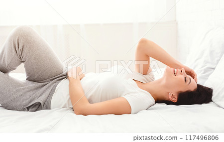 A young woman is lying comfortably on a bed with white linens, wearing casual grey sweatpants and a white t-shirt. She appears relaxed and happy, enjoying the sunny morning light A young woman is lying comfortably on a bed with white linens, wearing casual grey sweatpants and a white t-shirt. She appears relaxed and happy, enjoying the sunny morning light 117496806