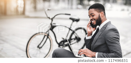 Black man professional is using his laptop and talking on his phone, enjoying an outdoor workspace with his bicycle next to him. The setting is dynamic and relaxed, capturing a modern work lifestyle. 117496831