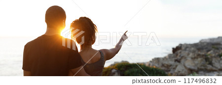 Young african american woman hugging man in sportswear, enjoy workout break, point finger at empty space in morning on rocks at ocean, outdoor. Relationship, health care, active lifestyle and sports 117496832