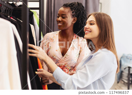 Two multiracial women examining various clothes on a rack in a bustling department store, both engrossed in their selections. Two multiracial women examining various clothes on a rack in a bustling department store, both engrossed in their selections. 117496837