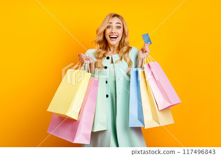 A woman wearing a light green dress stands against a bright yellow background, holding multiple colorful shopping bags and a credit card. She is smiling and looking directly at the camera A woman wearing a light green dress stands against a bright yellow background, holding multiple colorful shopping bags and a credit card. She is smiling and looking directly at the camera 117496843
