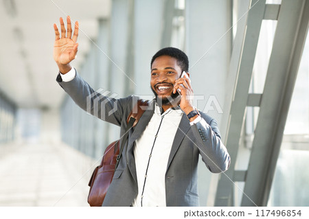 An African American businessman with a suitcase, talking on his phone outside an airport terminal. The scene is dynamic and professional, highlighting the fast-paced life of business travel. 117496854