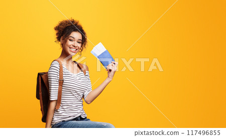 African American woman is seated on a chair, holding passport, flight tickets, isolated on yellow studio background, copy space 117496855