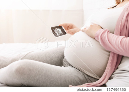 A pregnant woman is resting on a bed, comfortably dressed in casual clothing, while holding an ultrasound picture of her baby. Soft natural light filters through a window in the background, cropped 117496860