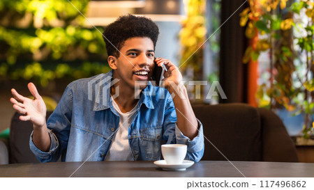 A young black guy is seated at a table, engaged in a phone conversation. He appears focused as he speaks on the phone. 117496862