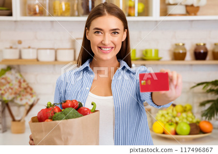 A woman stands in her kitchen, smiling and holding a grocery bag filled with produce and a credit card. 117496867