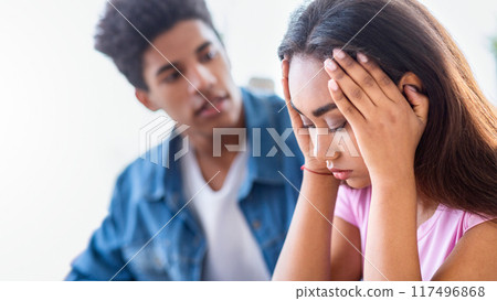 African American teen guy, wearing a blue denim shirt, looks at a teen girl with concern. The girl is seated with her hands covering her face, expressing sadness and distress. 117496868