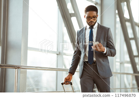 Black man with a suitcase is chatting on his phone outside an airport terminal. The scene is professional and busy, capturing the essence of corporate travel. 117496870