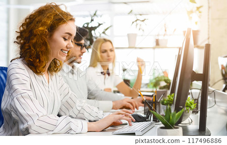 Several coworkers are seated at a desk, each engrossed in their computer screens, actively typing and clicking. The group appears focused and concentrated on their tasks 117496886