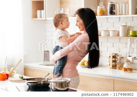 A woman is standing in a kitchen, holding a young child in her arms. The kitchen is well-lit with overhead lighting, and various cooking utensils can be seen in the background A woman is standing in a kitchen, holding a young child in her arms. The kitchen is well-lit with overhead lighting, and various cooking utensils can be seen in the background 117496902