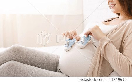 A pregnant woman is seated on a couch, holding her shoes in her hands. She appears to be taking a moment to rest and perhaps contemplating her day, cropped A pregnant woman is seated on a couch, holding her shoes in her hands. She appears to be taking a moment to rest and perhaps contemplating her day, cropped 117496903