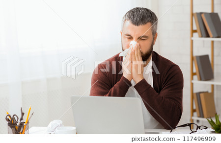 Sick man sitting in front of a laptop is blowing his nose into a tissue. He appears to be dealing with a cold or allergies while using the computer. Sick man sitting in front of a laptop is blowing his nose into a tissue. He appears to be dealing with a cold or allergies while using the computer. 117496909