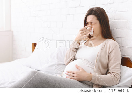 A pregnant woman sits on a bed in a well-lit, white-walled room. She covers her nose with a tissue and appears to be sneezing or dealing with a cold. A pregnant woman sits on a bed in a well-lit, white-walled room. She covers her nose with a tissue and appears to be sneezing or dealing with a cold. 117496948