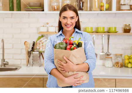 A woman stands in her kitchen, smiling, and holding a brown paper bag filled with fresh produce. A woman stands in her kitchen, smiling, and holding a brown paper bag filled with fresh produce. 117496986