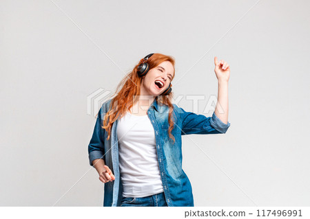 A jubilant young woman with red hair is lost in the joy of music, wearing headphones, her mouth open in mid-song and eyes closed. 117496991