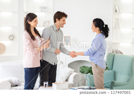 A young couple stands in a modern, well-lit living room, engaging with a real estate agent. The agent extends her hand in a professional handshake as they discuss potential property options. A young couple stands in a modern, well-lit living room, engaging with a real estate agent. The agent extends her hand in a professional handshake as they discuss potential property options. 117497001