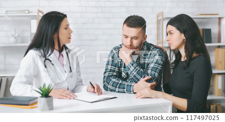 A doctor in a white coat is talking to a couple during a medical consultation in an office. The man is sitting with his head in his hands, while the woman is sitting next to him A doctor in a white coat is talking to a couple during a medical consultation in an office. The man is sitting with his head in his hands, while the woman is sitting next to him 117497025