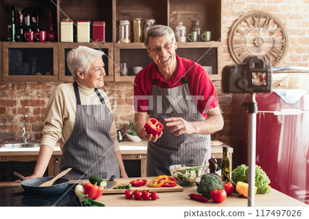 An older couple, wearing aprons, stand in their kitchen, smiling and holding vegetables, as they film a cooking video with a camera mounted on a tripod. 117497026