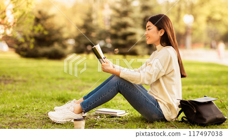 Asian woman is sitting on the grass, engrossed in a book she is reading. The sunlight highlights her features as she enjoys her reading time outdoors, side view 117497038