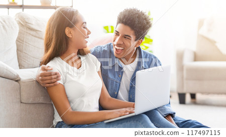 African American teen couple sits together on a light-colored floor inside a home. The guy is using a laptop, and both are laughing. 117497115