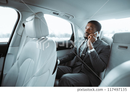 African American businessman in a gray suit is sitting in the back seat of a car and talking on a cell phone. He is looking out the window with a thoughtful expression on his face. 117497135