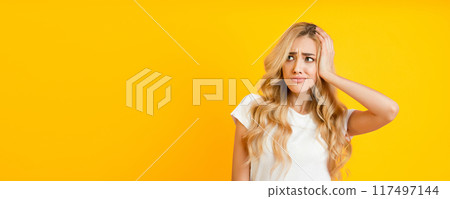 A young woman with long blonde hair is standing against a yellow background, looking off to the side with a confused expression. She is wearing a white t-shirt and has one hand on her head. 117497144