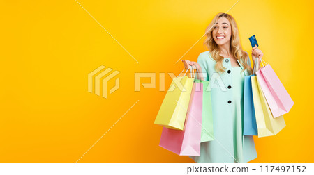 A happy woman with long blonde hair is standing in front of a yellow background. She is holding several shopping bags in one hand and a credit card in the other hand, copy space A happy woman with long blonde hair is standing in front of a yellow background. She is holding several shopping bags in one hand and a credit card in the other hand, copy space 117497152