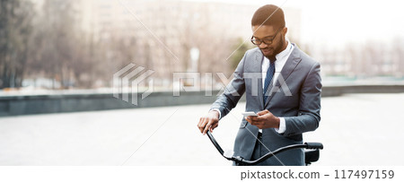 Black businessman on a bicycle is in the city, checking his smartphone. The scene is energetic and contemporary, highlighting a seamless blend of work and transportation in an urban setting. 117497159