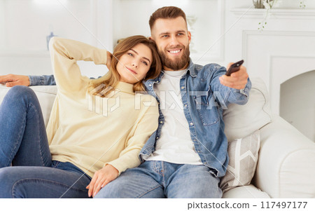 A cheerful couple sits on a comfortable sofa, enjoying their time watching television together in a warm, well-decorated living room. 117497177