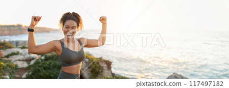 African American young woman in athletic wear stands on a rocky cliff overlooking the ocean. She has her arms raised in the air, fists clenched, and is smiling with excitement, copy space African American young woman in athletic wear stands on a rocky cliff overlooking the ocean. She has her arms raised in the air, fists clenched, and is smiling with excitement, copy space 117497182