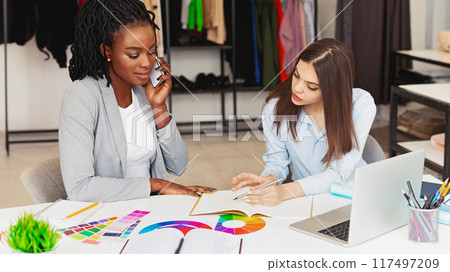A focused scene unfolds with two professional multiracial women working closely together at a white table filled with vibrant color samples. A focused scene unfolds with two professional multiracial women working closely together at a white table filled with vibrant color samples. 117497209