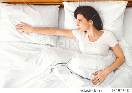 A woman lying in bed wearing white pajamas reaches out to an empty pillow next to her. The scene is captured in a brightly lit bedroom during the morning. A woman lying in bed wearing white pajamas reaches out to an empty pillow next to her. The scene is captured in a brightly lit bedroom during the morning. 117497218