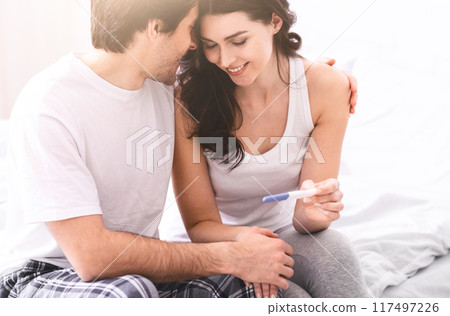 A young couple is sitting on a bed, the woman is holding a pregnancy test in her hand while the man puts his arm around her shoulder. Both are smiling and looking at the test. 117497226