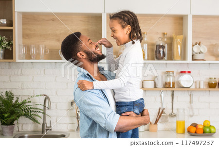 Little african girl and her cheerful father bonding at kitchen, daughter playing with dad nouse, empty space Little african girl and her cheerful father bonding at kitchen, daughter playing with dad nouse, empty space 117497237